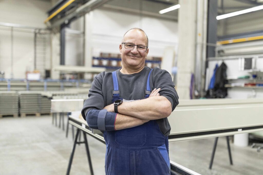 Ein Arbeiter in blauer Latzhose steht laechelnd in einer Werkstatt.