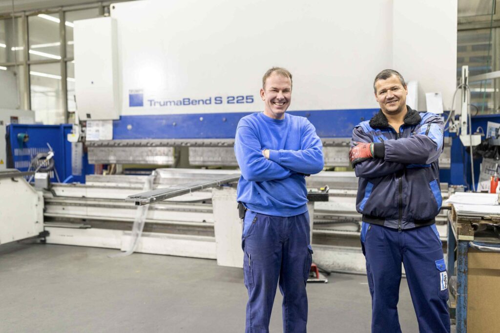 Zwei Maenner stehen vor einer großen Maschine in einer Fabrikhalle.